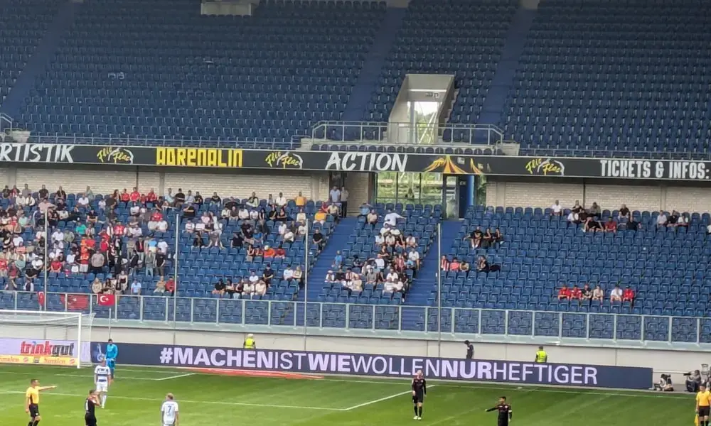 Show thumbnail preview Ein Fußballspiel in einem teilweise gefüllten Stadion. Spieler sind auf dem Feld, während ein digitales Banner Text auf Deutsch anzeigt. Die Zuschauer sind über die Sitze verteilt.