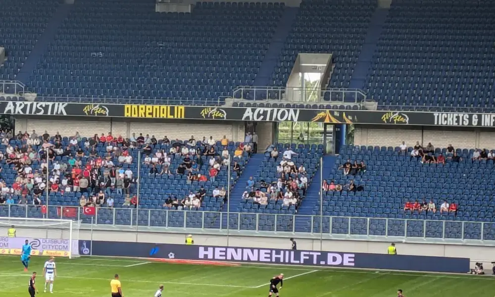 Ein spärlich besuchtes Fußballspiel in einem großen Stadion mit blauen Sitzen. Im Hintergrund sind die Anzeigetafel und Werbung zu sehen, und auf dem Feld sind einige Spieler. Datenschutzerklärung