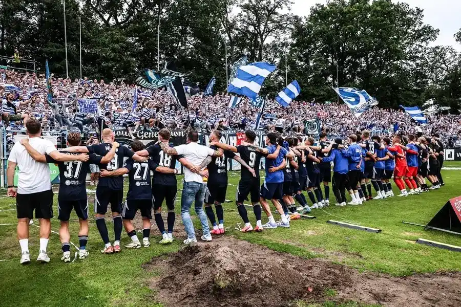 Eine Fußballmannschaft steht Arm in Arm vor jubelnden Fans und hält blau-weiße Fahnen in einem Stadion.