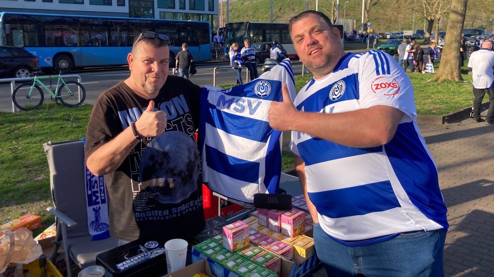 Zwei MSV-Fans präsentieren ein Trikot am Stadion-Vorplatz, umgeben von Verpflegung.