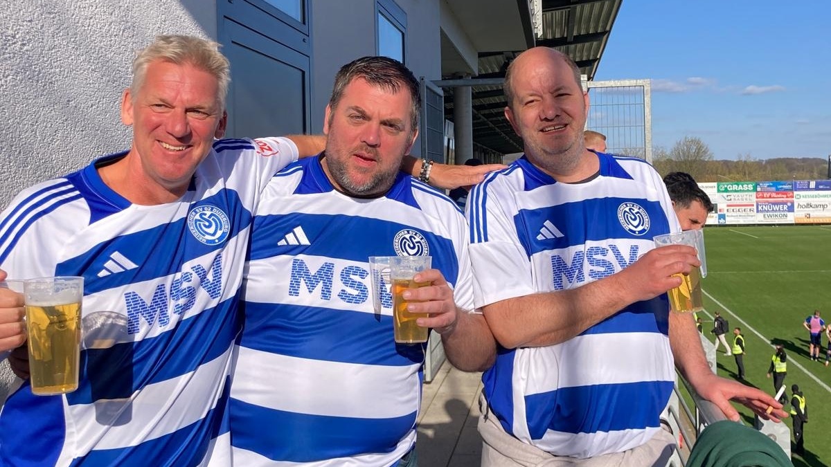 Drei MSV-Fans mit Trikots und Bier auf einer Stadionterrasse bei Sonnenschein.