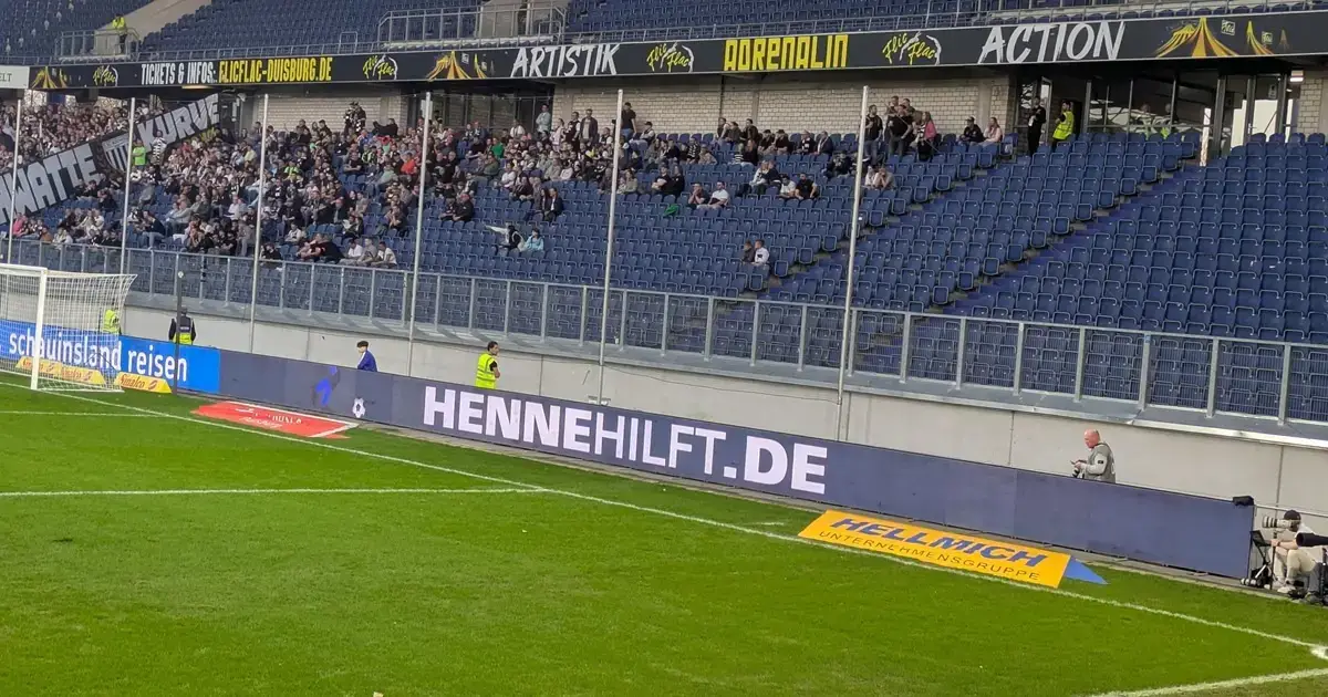 Rückblick Werbebande mit der Aufschrift Henne hilft im Stadion neben dem Torbereich und Zuschauerrängen.