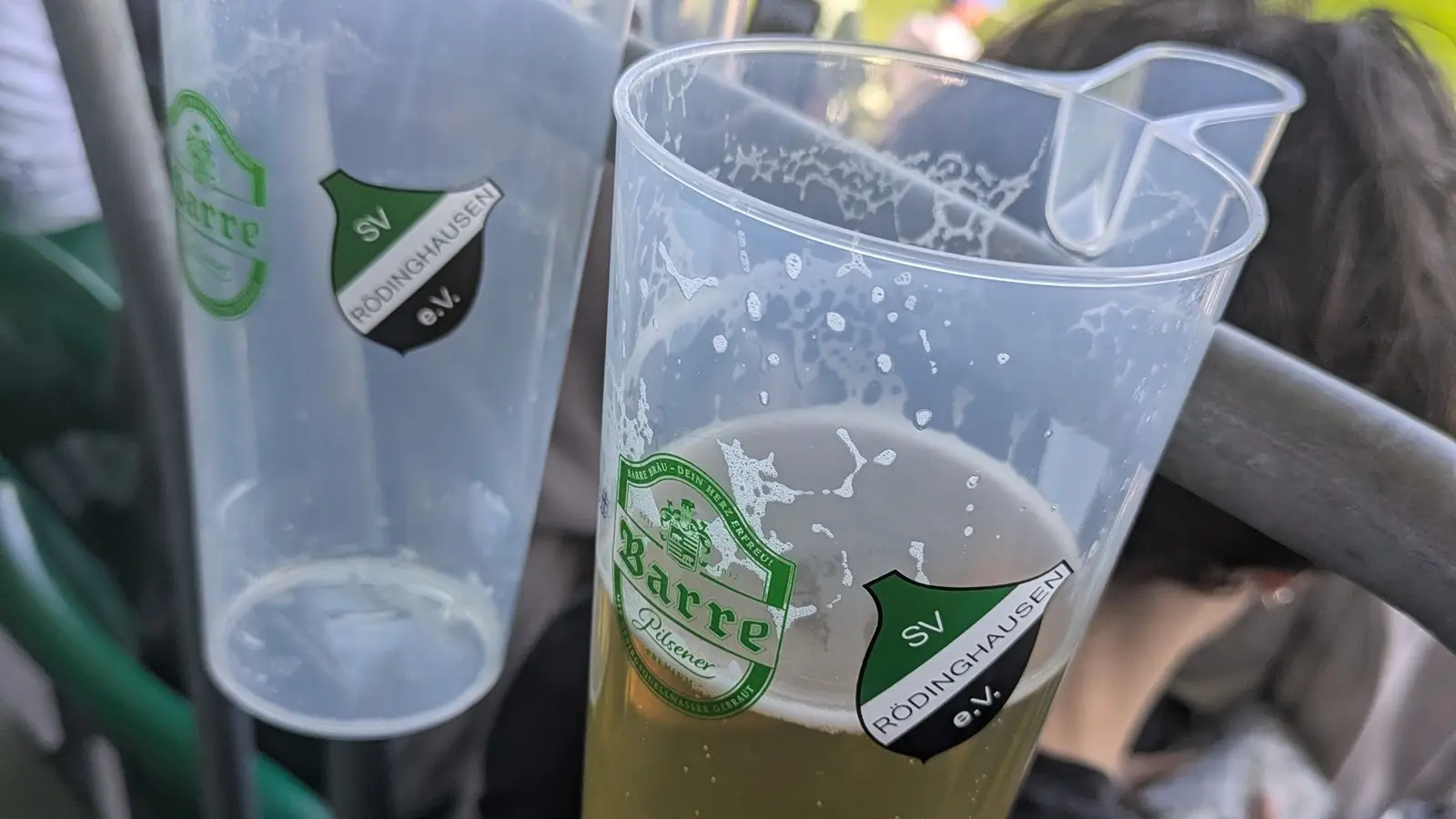 Zwei Bierbecher mit Logos des SV Rödinghausen und Barre Pilsener auf Stadiongeländer.